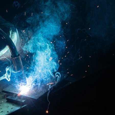 Close-up of a pipe fitting being welded to a steel baseplate, weld pool and shielding gas smoke visible